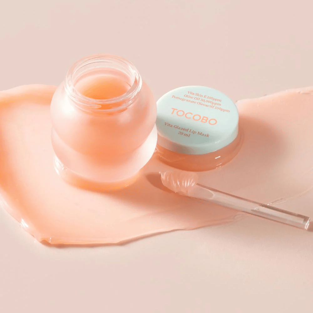 Een pot met "Vita Glazed Lip Mask" van Tocobo wordt getoond. De pot is doorzichtig met een licht turquoise deksel en bevat een glanzend, roze lipmasker. Een kleine spatel ligt naast de pot en een deel van het masker is uitgespreid op het oppervlak. Het product is ontworpen om de lippen te hydrateren en te verzorgen.