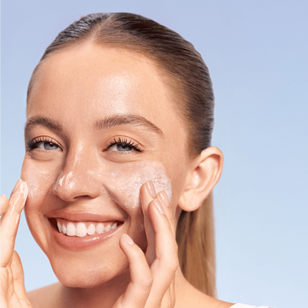 Lachende vrouw brengt LANEIGE Water Bank Gentle Gel Cleanser in schuimende vorm aan op de wangen, tegen blauwe achtergrond.