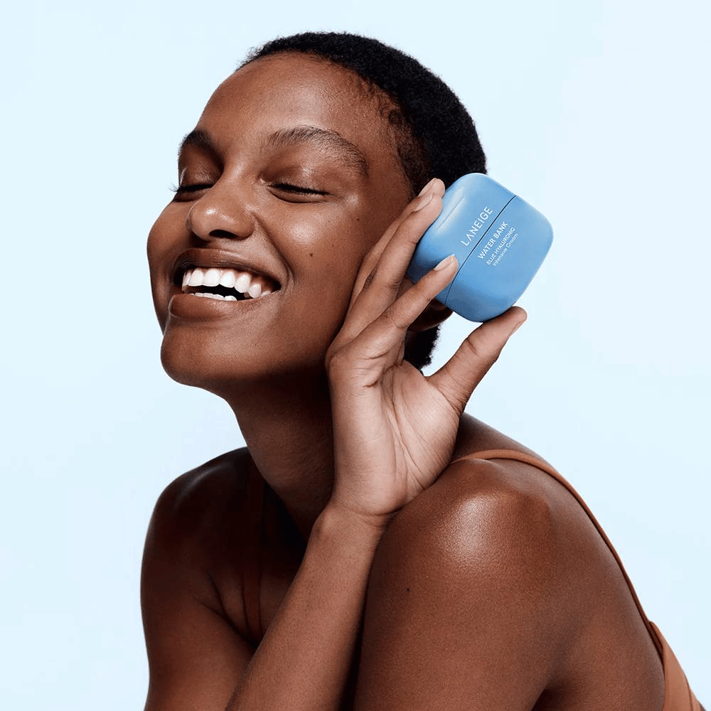 Lachende vrouw houdt een blauwe pot LANEIGE Water Bank Blue Hyaluronic Cream tegen haar wang, met een lichtblauwe achtergrond.