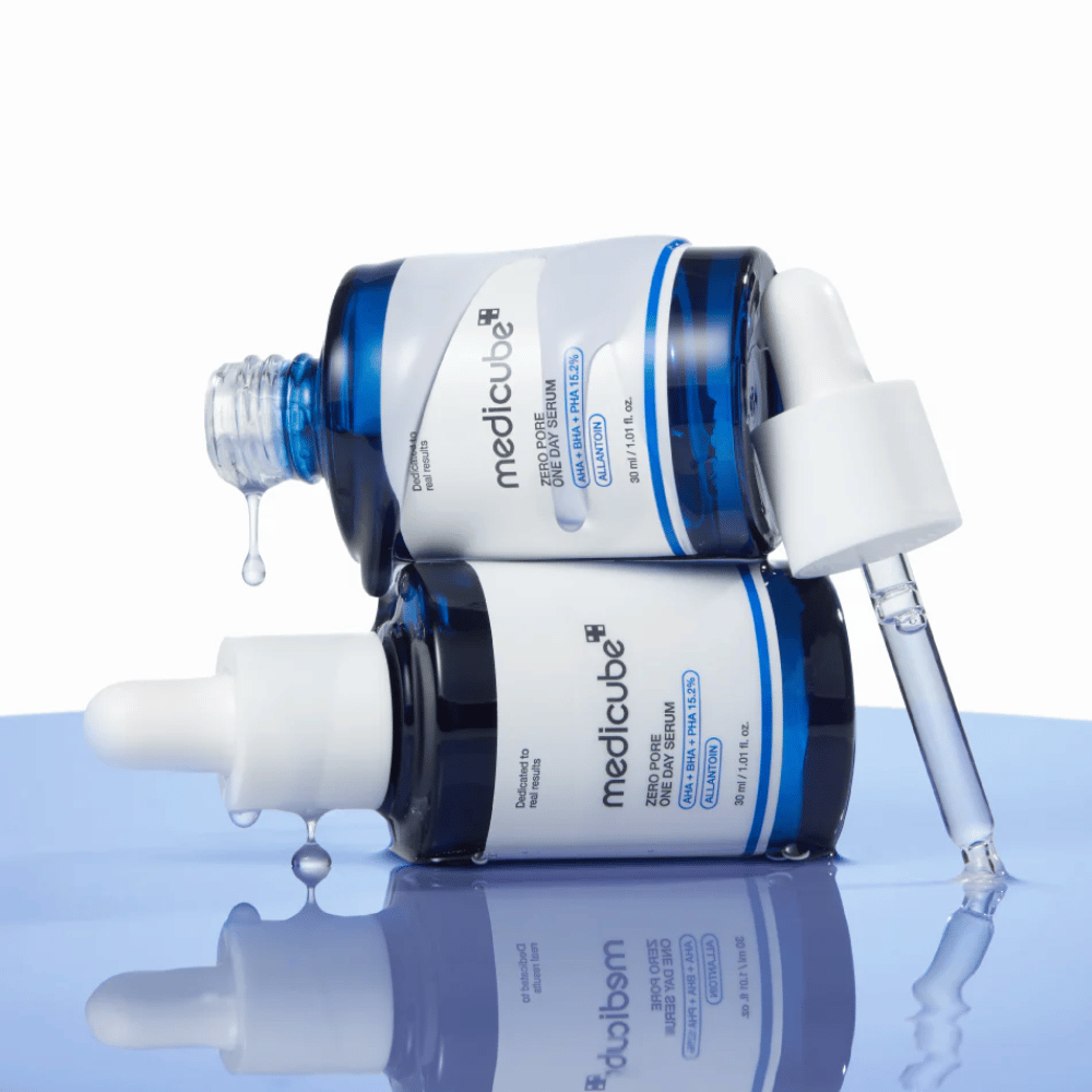 Twee blauwe flessen van Medicube Zero Pore One Day Serum liggend tegen elkaar, met pipetten en druppels serum op een reflecterend blauw oppervlak.