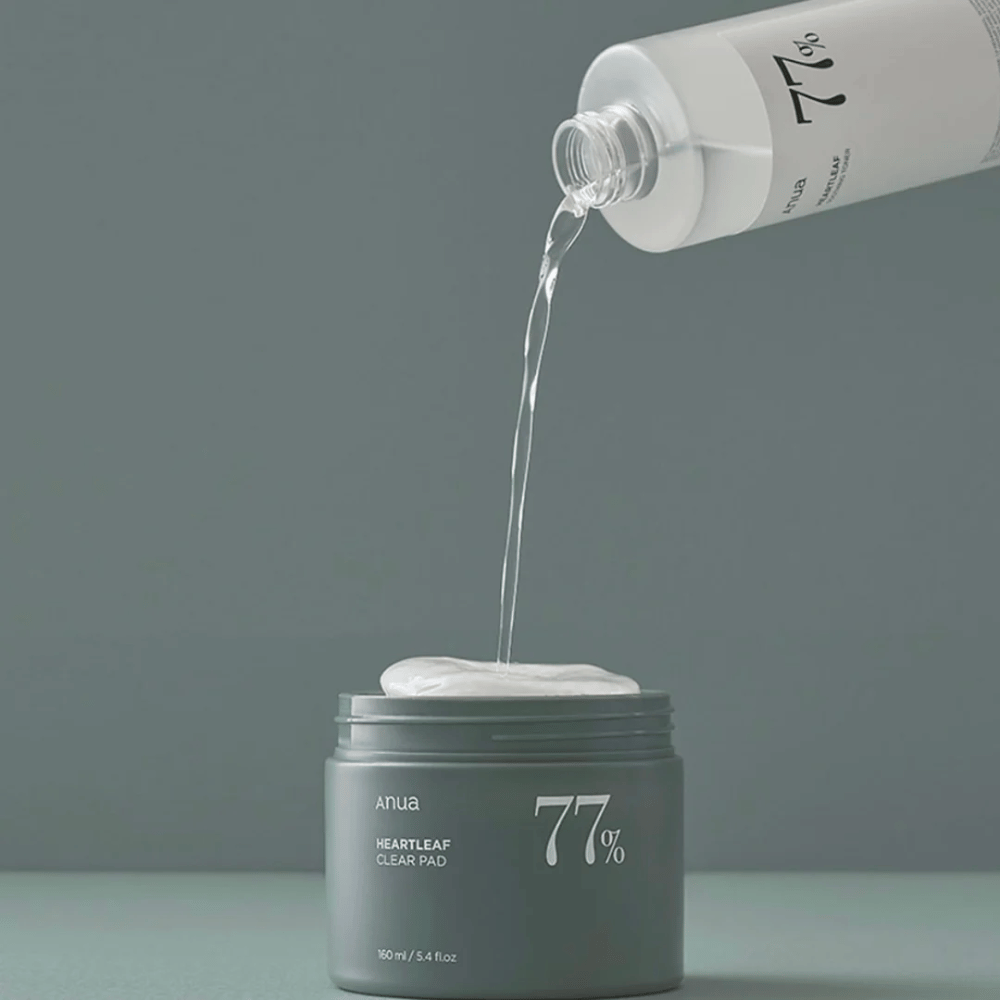 Een doorzichtige fles giet een vloeistof in een grijze pot gemarkeerd met "anua HEARTLEAF 77% Clear Pad", die een stapel witte huidverzorgingspads bevat. De achtergrond is eenvoudig en effen in een zachte grijze tint.