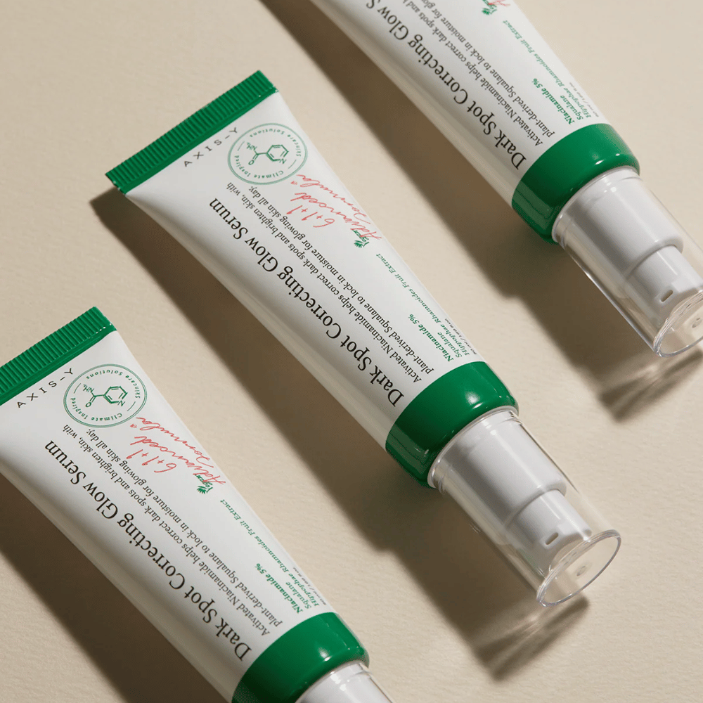 Drie tubes van Axis-Y Dark Spot Correcting Glow Serum met groene dop en witte verpakking liggen op een lichte ondergrond.