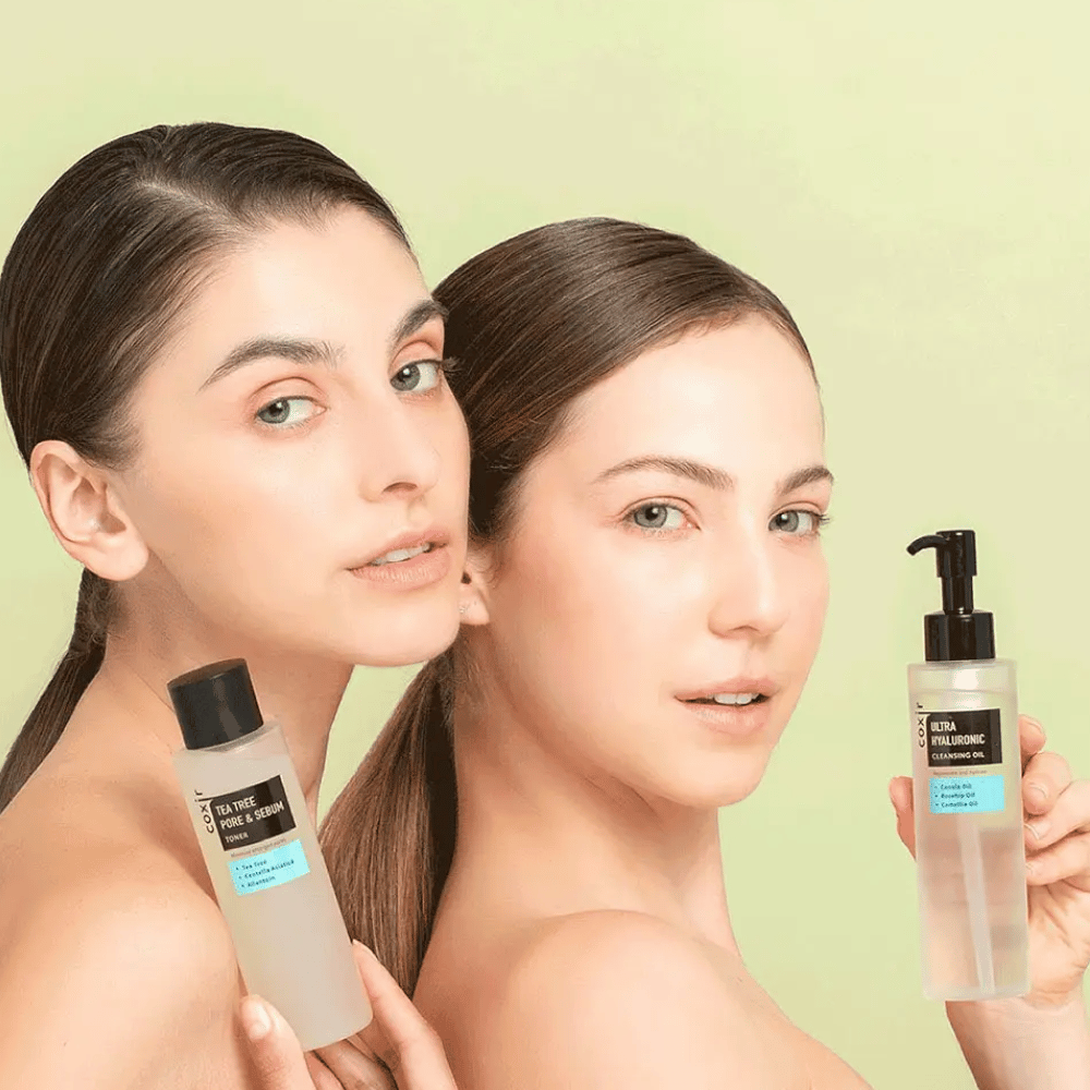 Twee vrouwen met ontblote schouders poseren met huidverzorgingsproducten; de ene houdt een fles vast met het label "TEA TREE PORE & SEBUM", en de andere een pompflacon met het label "ULTRA HYALURONIC CLEANSING OIL". Ze staan voor een lichtgroene achtergrond.