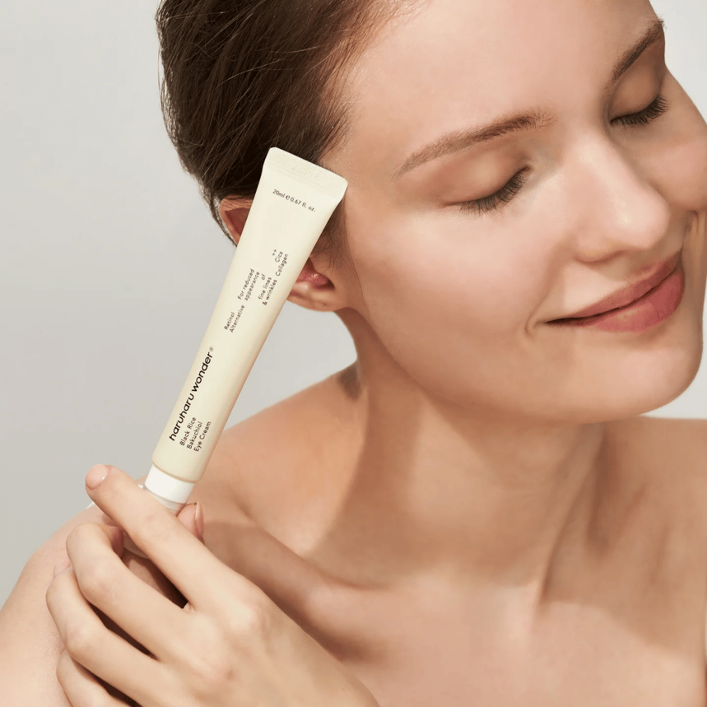 Een glimlachend persoon met gesloten ogen houdt een tube Haruharu Wonder Black Rice Bakuchiol Eye Cream tegen de slaap.