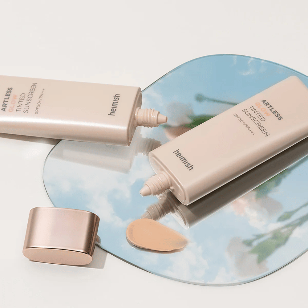 Een fles heimish Artless Glow Tinted Sunscreen SPF50+ PA+++ 40ml en een fles concealer op een spiegel, die verschillende huidtinten toont.