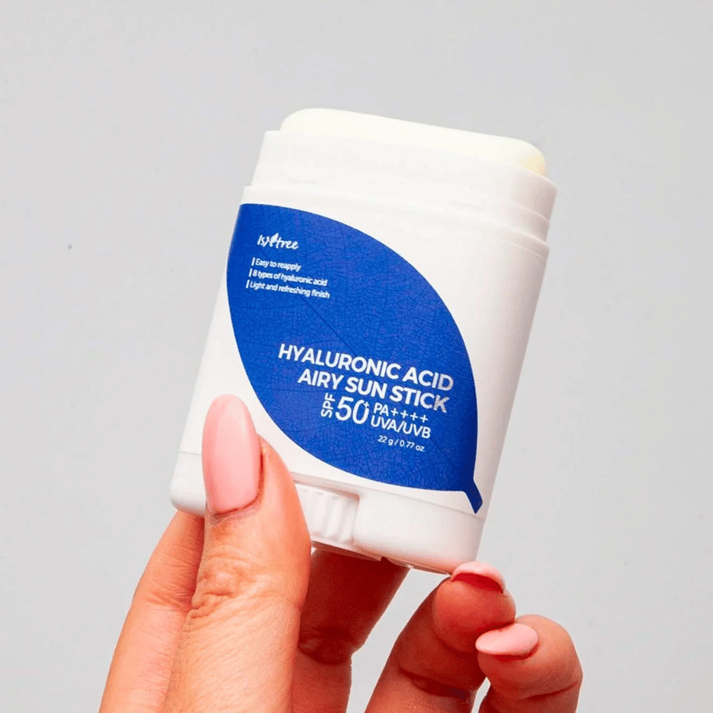 Een hand houdt een witte zonnebrandstick met blauw etiket van het merk Isntree. Het product heet "Hyaluronic Acid Airy Sun Stick" en heeft een zonbeschermingsfactor van SPF 50+ PA++++.