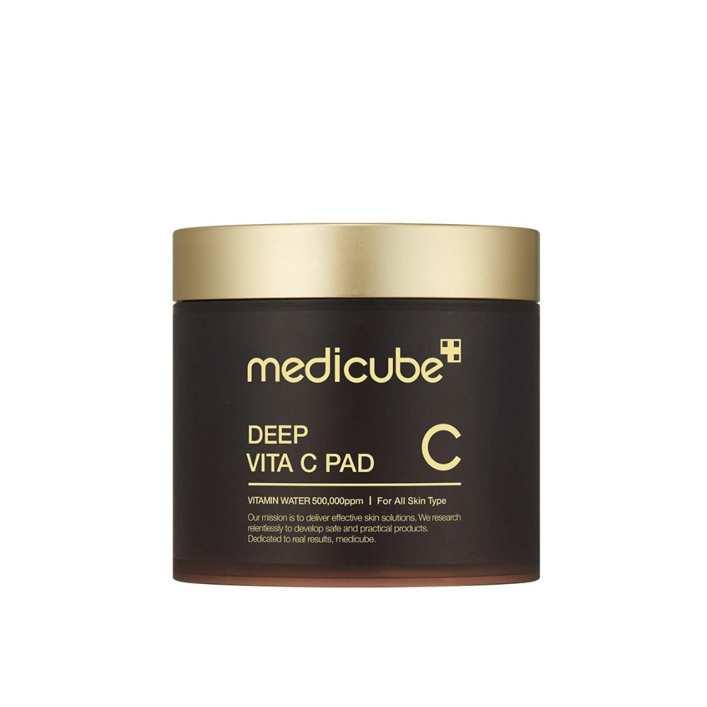 Een pot van medicube, gemarkeerd met "Deep Vita C Pad", met een gouden deksel en een donkere body. Tekst op de pot geeft aan dat het vitamine C bevat en geschikt is voor alle huidtypes. De achtergrond is neutraal.