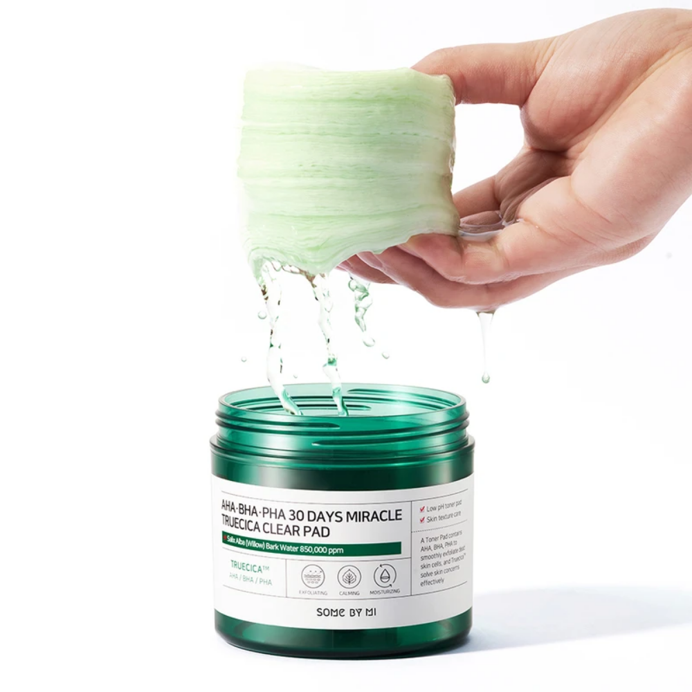 Een hand giet een groene vloeistof in een pot voor SOME BY MI:s AHA, BHA, PHA 30 Days Miracle Truecica Clear Pad 70 stuks acne-uitbraak behandeling.