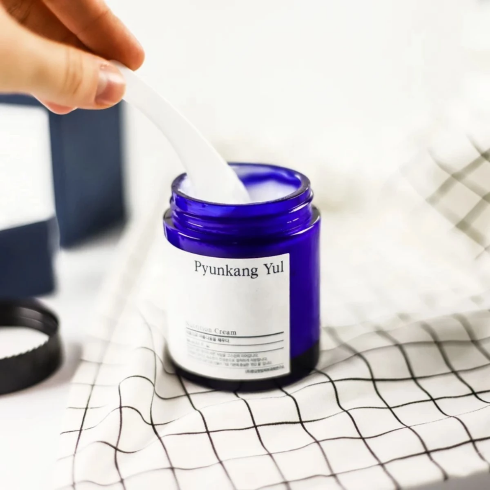 Een persoon gebruikt een pot Pyunkang Yul Nutrition Cream 100ml op een tafel.