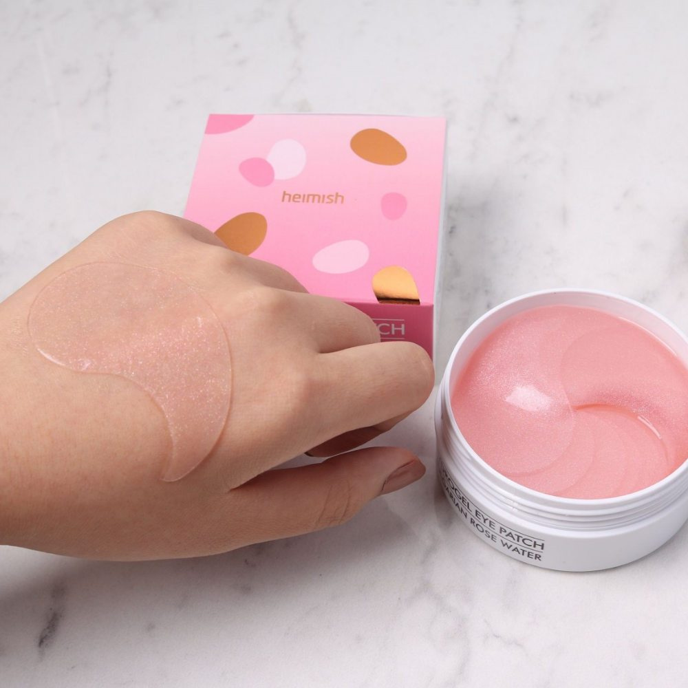 Een hand die een roze Bulgarian Rose Hydrogel Eye Patch 60st van heimish vasthoudt en een oogmasker aanbrengt.