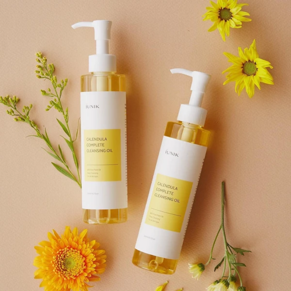 Twee flessen iUNIK Calendula Complete Cleansing Oil 200ml met gele bloemen op beige achtergrond, doordrenkt met plantaardige olie.