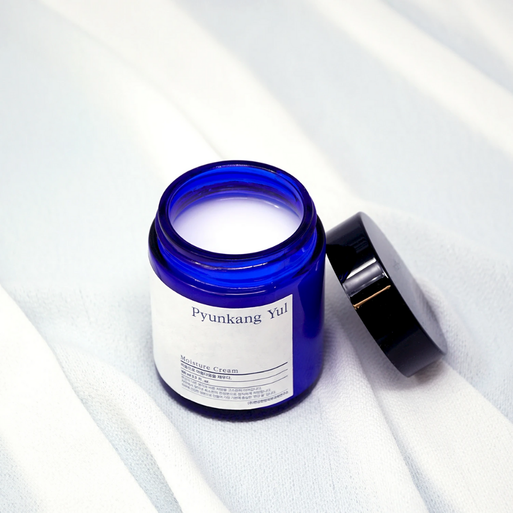 Een blauwe pot met deksel die op een witte doek zit en Pyunkang Yul's Moisture Cream 100ml hydraterende eigenschappen toont.