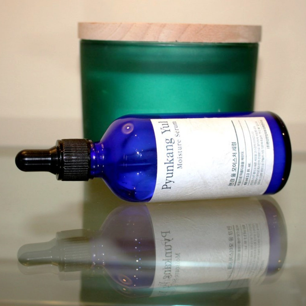 Een fles Pyunkang Yul Moisture Serum 100ml reguleert de vochtbalans op een tafel naast een kaars.